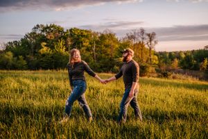 A couple walks hand in hand through a sunlit meadow during golden hour with trees in the background.