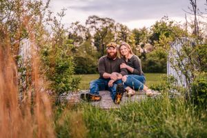 A couple sits together in a grassy field at sunset surrounded by lush greenery.
