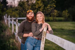 A couple poses together by a white fence with green fields and trees in the background.