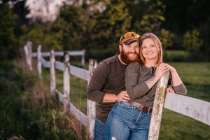 A casual couple leans against a white fence in a rustic countryside setting at sunset.