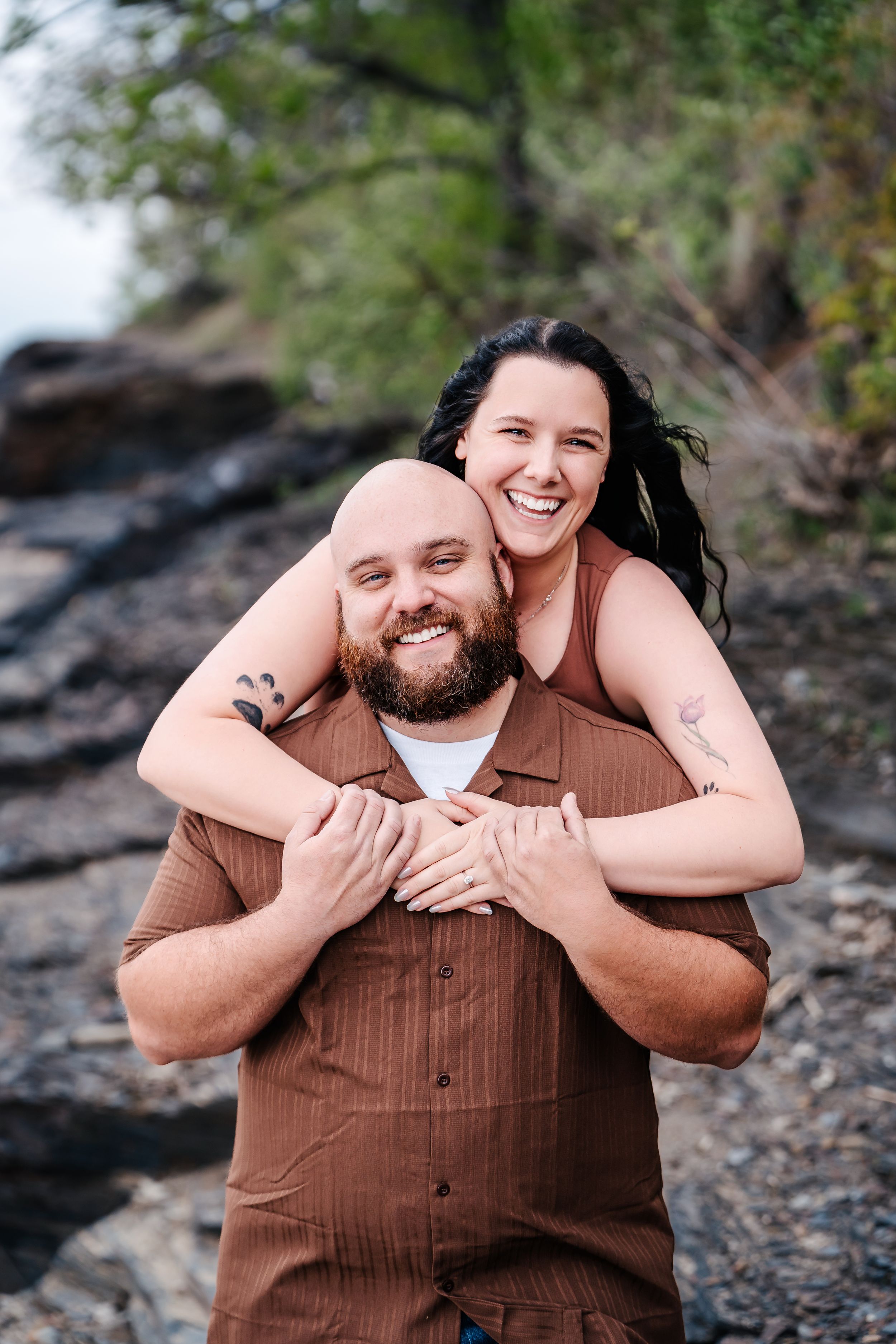A joyful couple embraces on rocky terrain with trees in the background during an outdoor photo session.