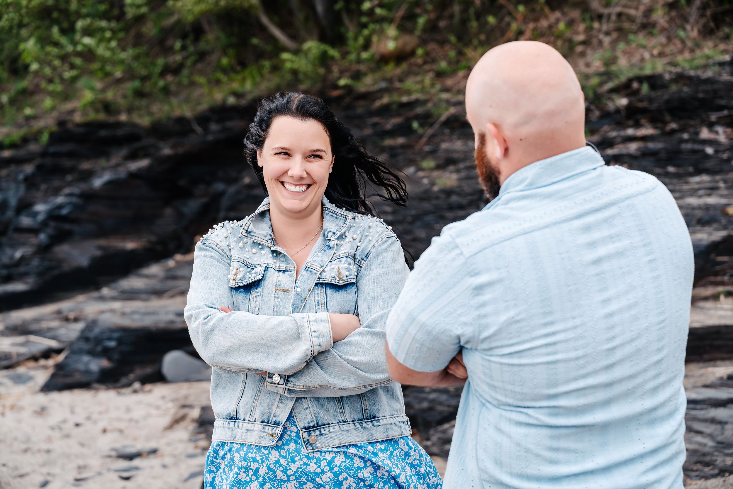 Two people having a cheerful conversation while sitting on rocks at a beach shore with rocky cliffs in the background.