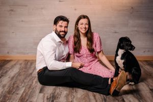 Couple sits on wooden floor with black and white dog in casual portrait setting.