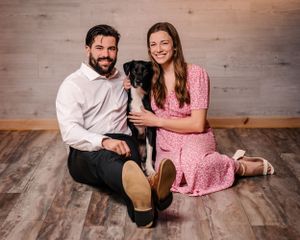 Couple in formal wear sits on hardwood floor with their black dog for family portrait.