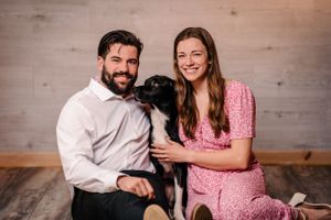 Indoor portrait with black dog sitting between couple on wooden floor.