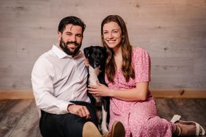 Couple in pink dress and white shirt poses with black dog against wooden wall.