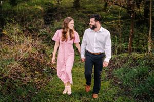 Couple walking hand in hand through a lush forest path wearing pink dress and white shirt.