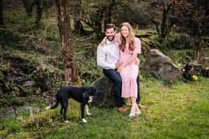 Couple poses with their black and white dog in a wooded outdoor setting.