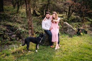 Happy couple poses with their black and white dog in a green forest setting.