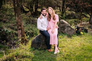 Couple in pink and white sits on a rock with forest scenery behind them.