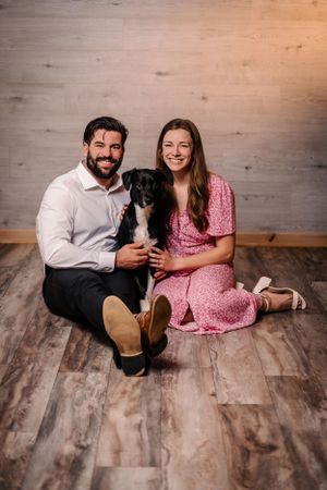 Family portrait with black puppy sitting between couple on wooden floor.