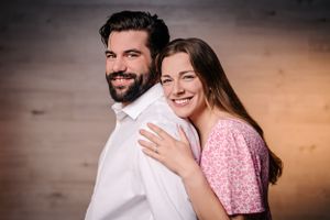 Couple smiles in side-by-side pose with dramatic lighting and wooden backdrop.