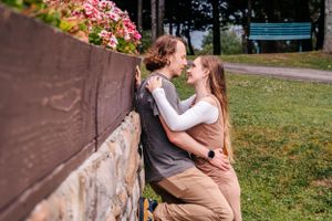 A romantic couple embraces against a stone wall with pink flowers blooming nearby in a garden setting.