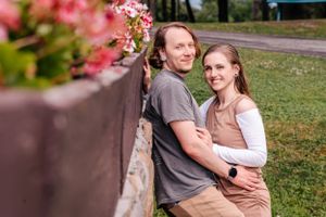 A couple poses together near a decorative wall with pink flowers in a park setting.