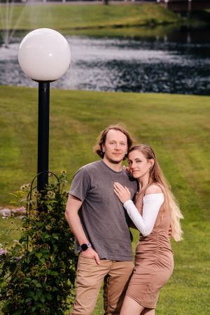 A couple poses near a white globe lamp post with a pond in the background.