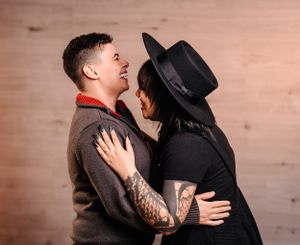 An intimate indoor portrait of a couple in dark clothing with one wearing a black hat.