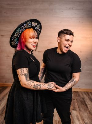 Two people in black outfits share a laugh while showing their tattooed arms.