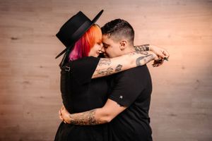 Tattooed couple in black outfits embracing intimately against warm-toned wooden backdrop.