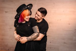 Couple in matching black outfits sharing romantic moment, one wearing decorative hat.