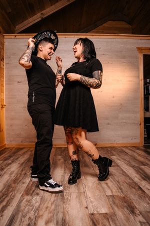 Alternative couple in black outfits dancing playfully on wooden floor.