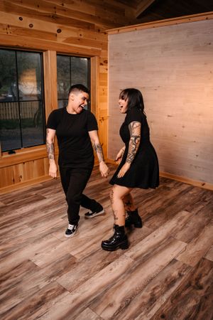 Playful tattooed couple dancing together on wooden floor in rustic cabin setting.