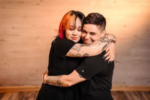 Intimate couple embrace in black outfits against wooden wall with warm lighting.