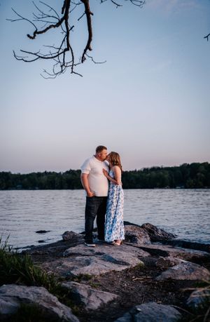 A romantic couple stands together on rocky lakeside terrain at dusk with tree branches visible overhead.