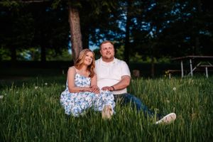 A couple sits together in tall grass during a summer photoshoot outdoors.