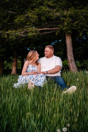 A couple in coordinated outfits share an intimate moment while sitting in a grassy field near pine trees.