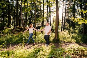 Playful couple runs through sunlit forest holding hands among tall pine trees.