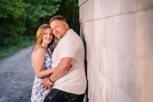 Couple embraces against white wall on rustic path surrounded by greenery.