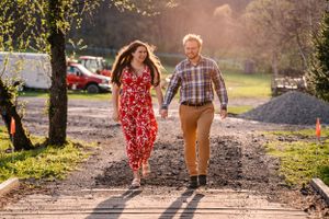 A couple walks hand in hand down a sunlit path wearing a red floral dress and plaid shirt.