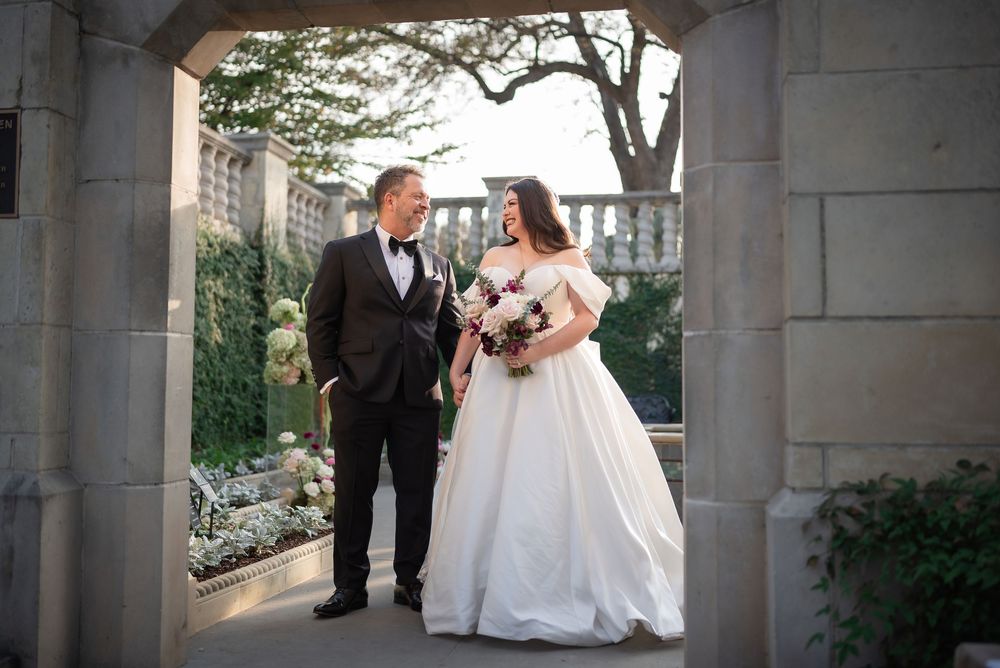 Elegant bride and groom walking through stone archway in garden during golden hour wedding portrait