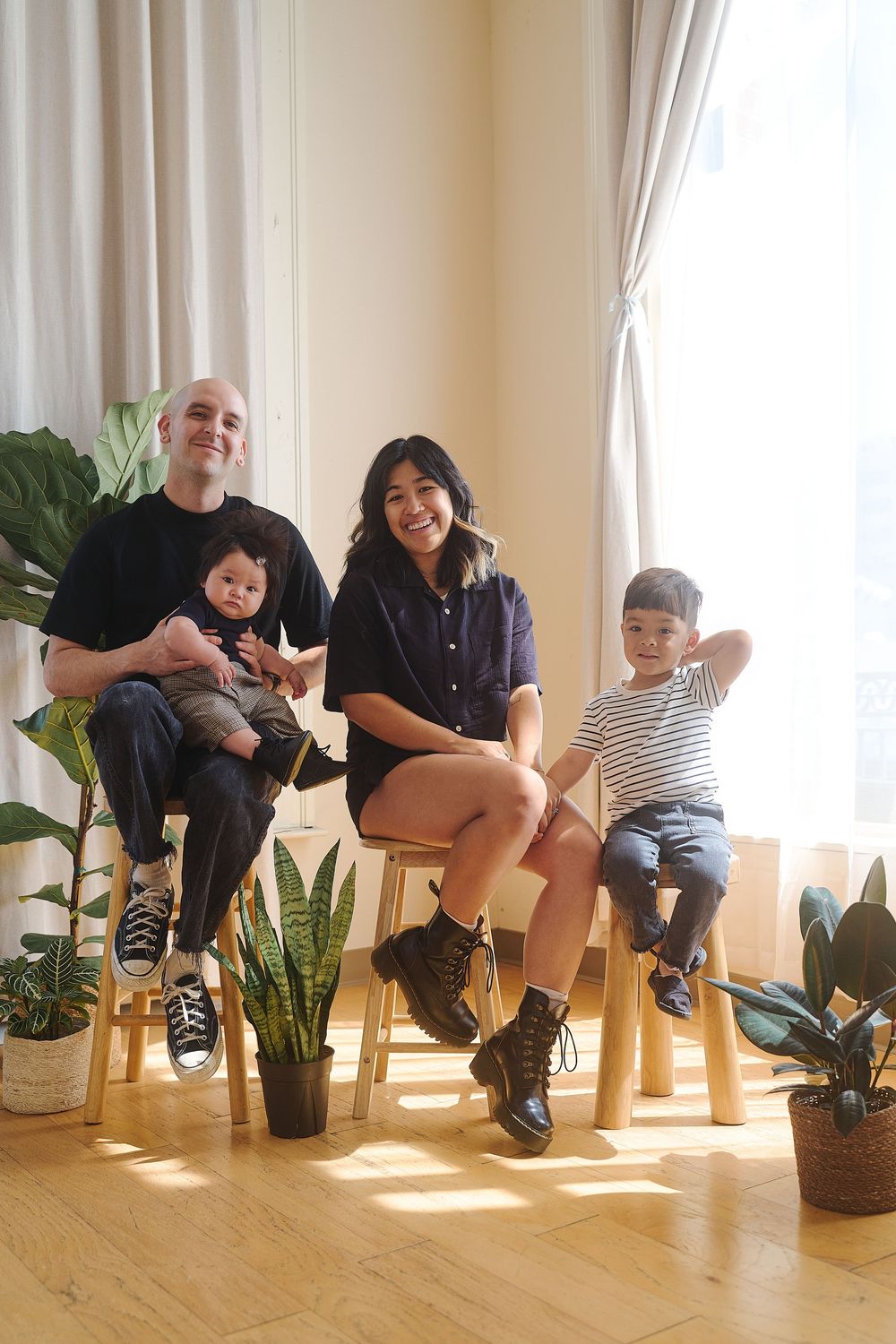 A happy interracial family of four sits together on stools surrounded by indoor plants in a bright room.