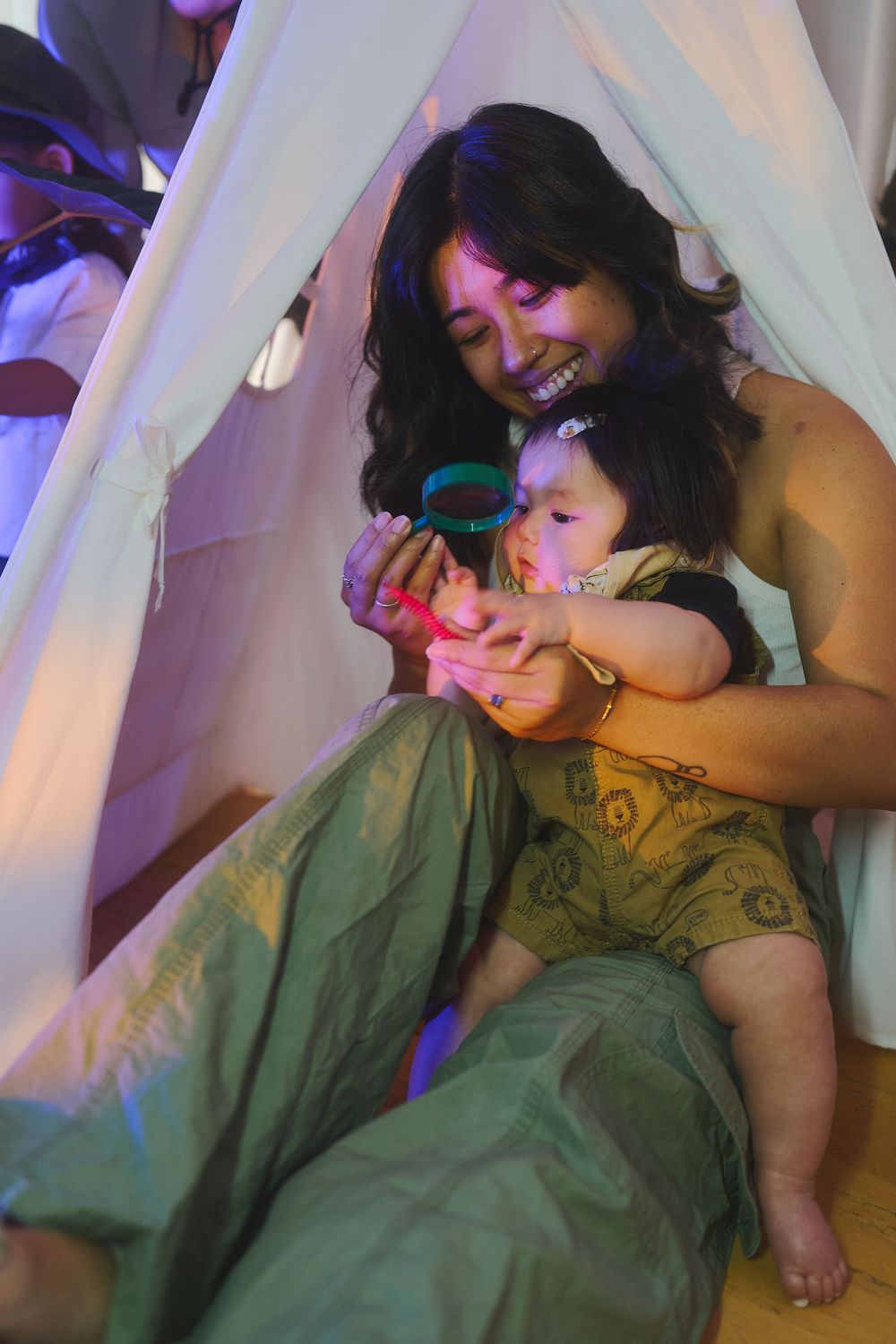 A mother and baby play with colorful toys inside a glowing teepee tent with purple and pink lighting.