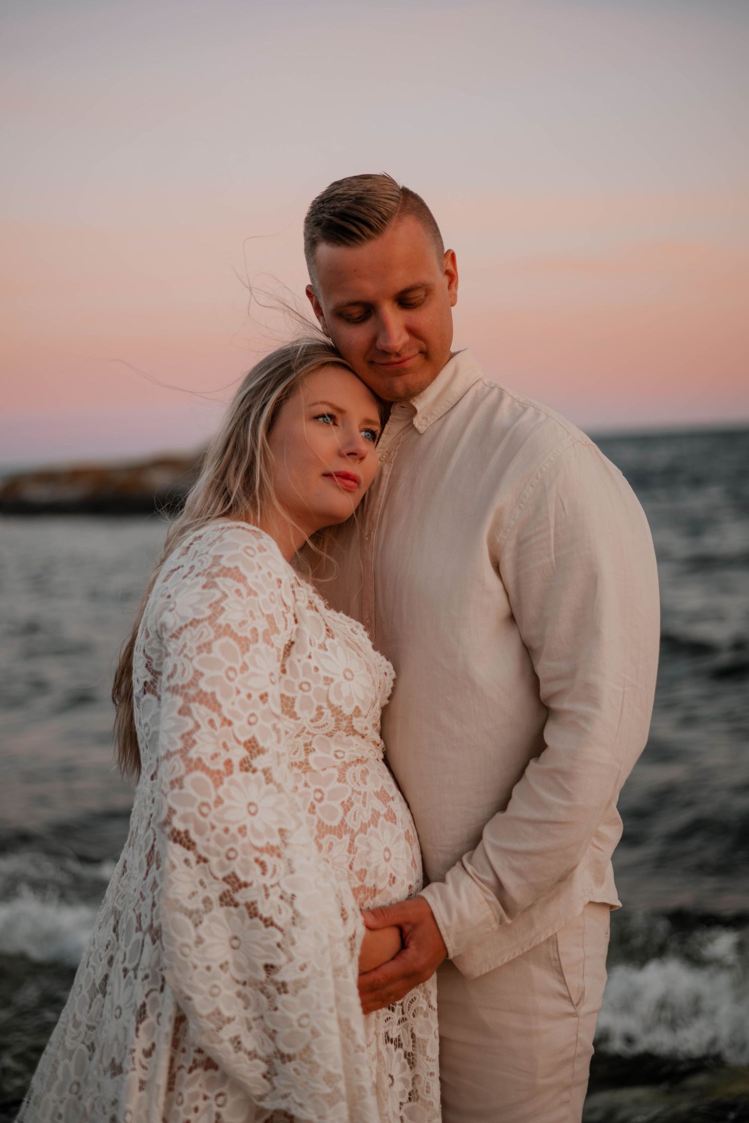 Ett gravidpar står på en klippa vid havet under rosa himmel