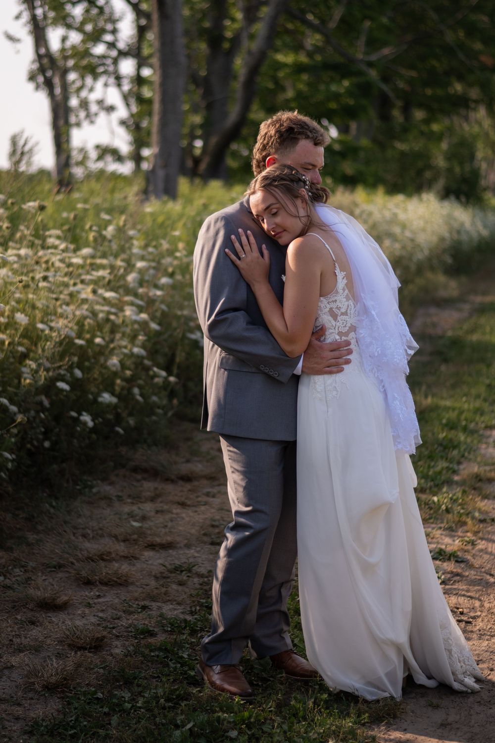 A couple shares an intimate moment in their wedding attire while standing outdoors in a wooded setting.