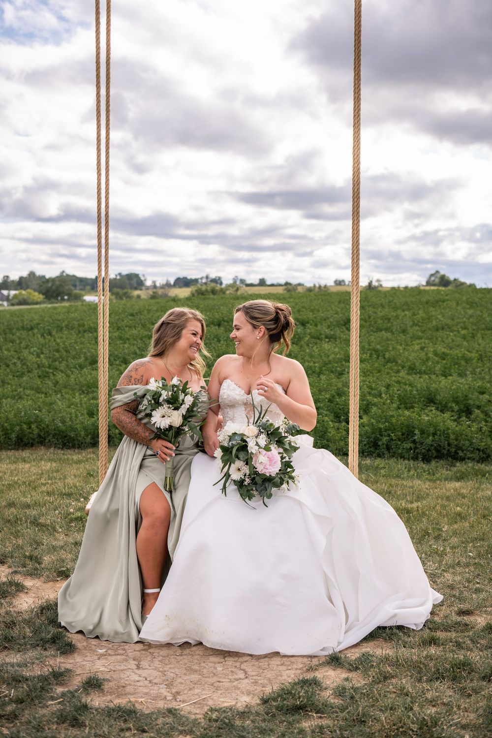 Two people in wedding dresses sit together on wooden swings in a rustic outdoor setting.