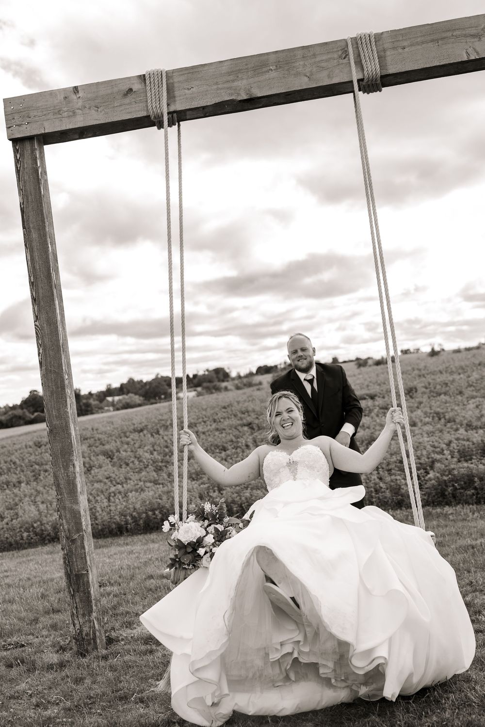 A romantic wedding couple sits together on a rustic wooden swing in a scenic countryside field.