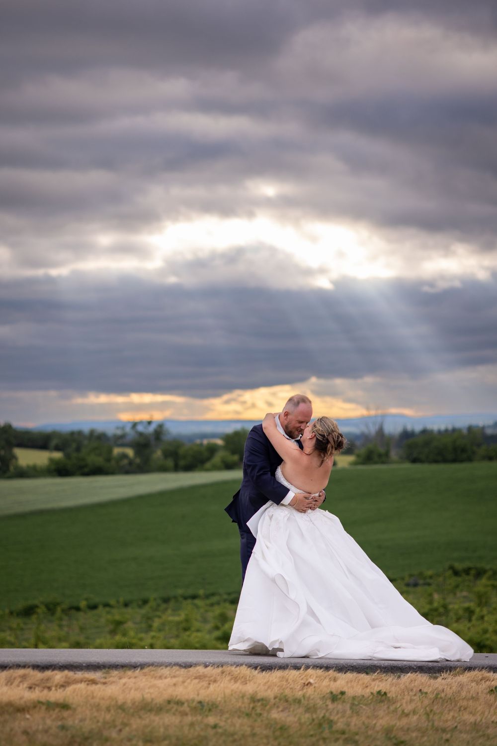 A couple in wedding attire shares a romantic moment on a hilltop overlooking green fields under dramatic cloudy skies.