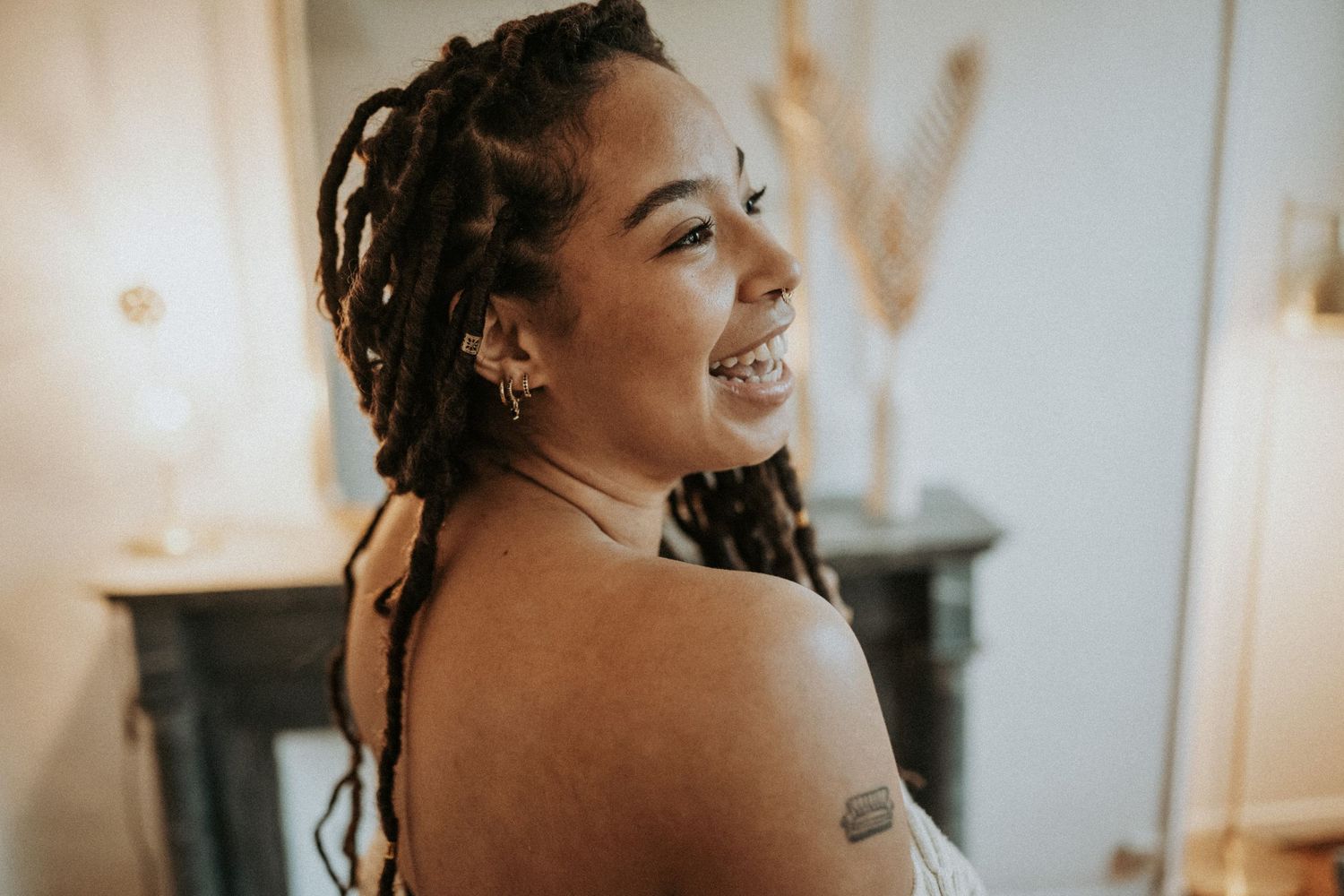 An intimate portrait captures a joyful moment with dreadlocks and bare shoulders against a light interior background.