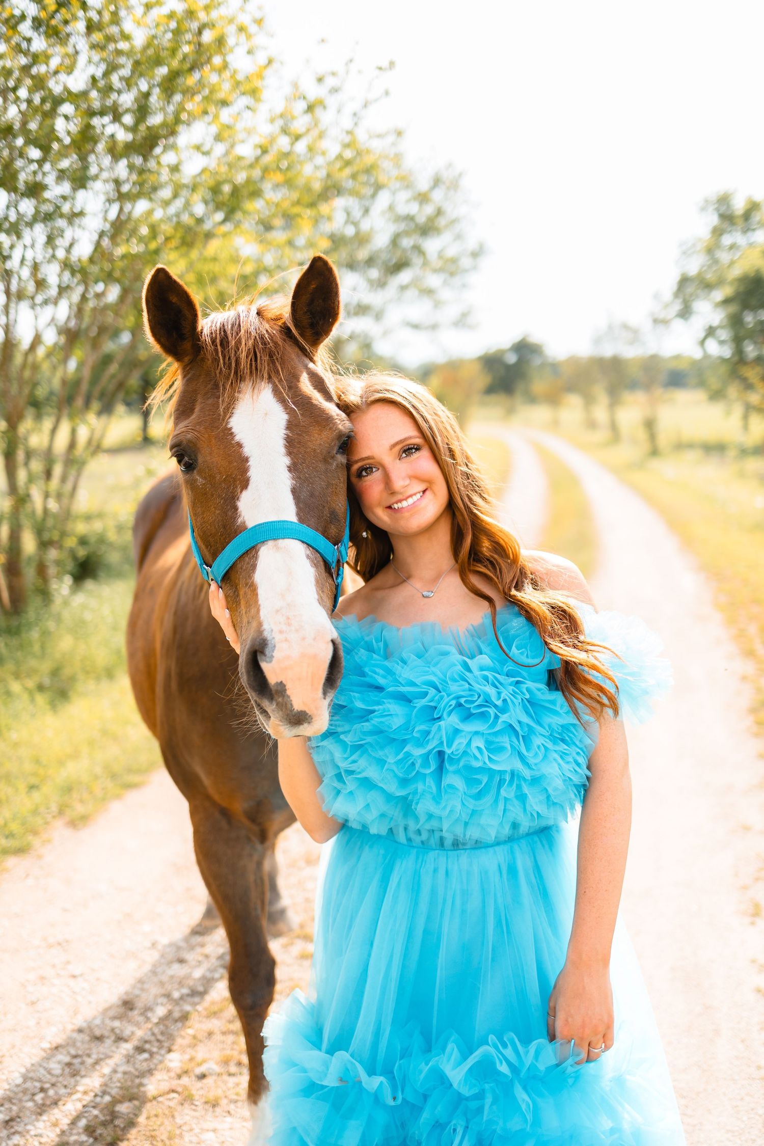Smiling young woman in blue ruffled dress poses with chestnut horse on sunny dirt road.