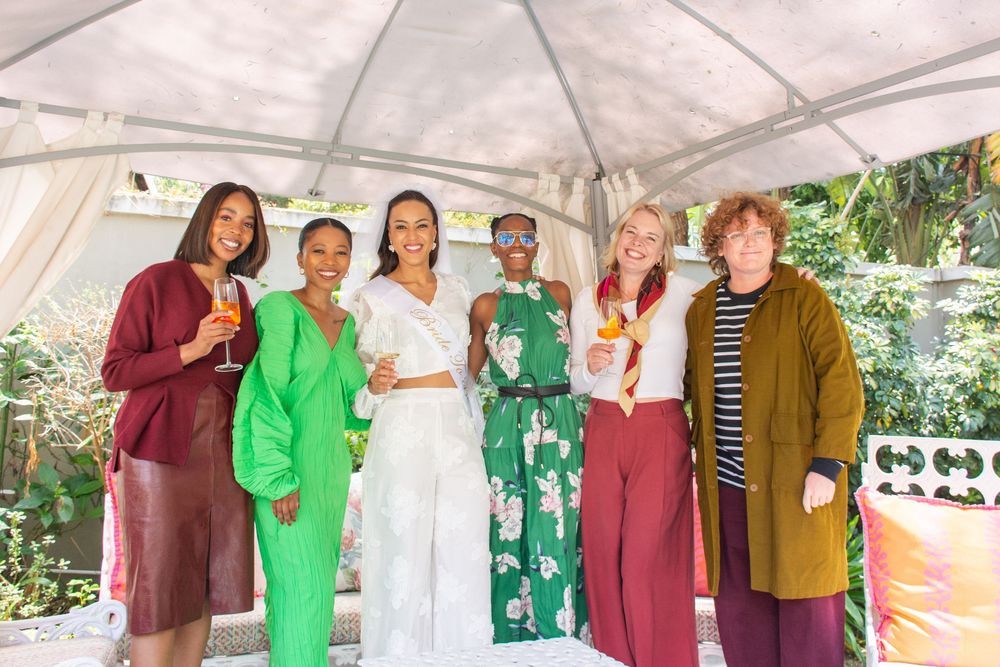 Group of friends in casual attire gathered outdoors at a social event with white tent and greenery in background.