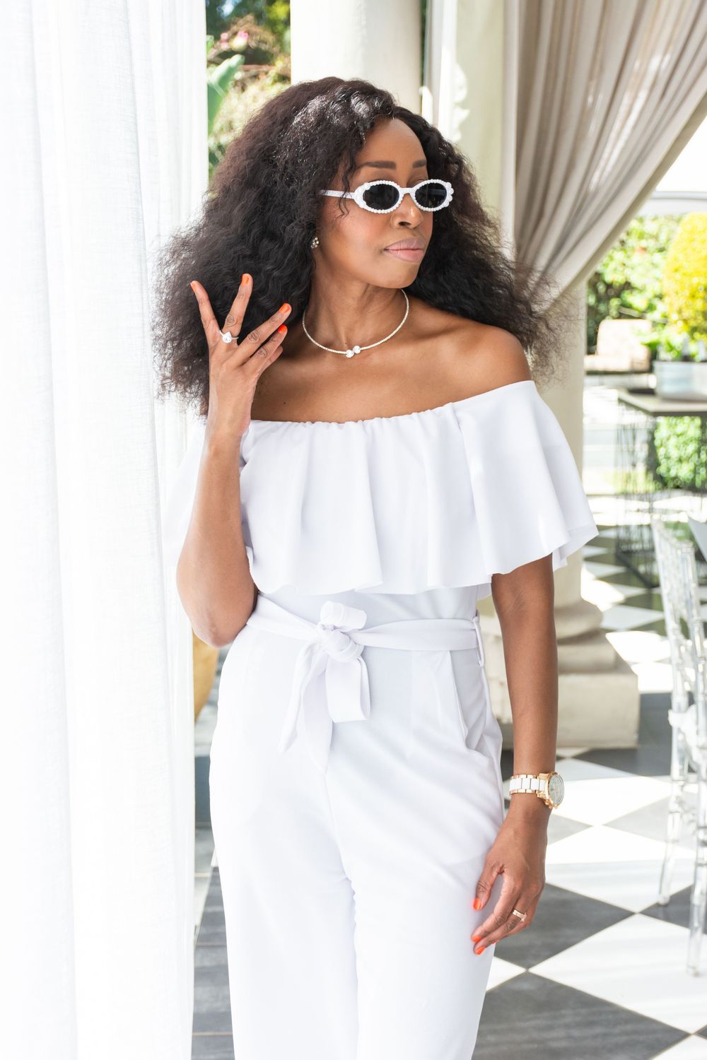 Woman wearing white off-shoulder summer dress and sunglasses poses elegantly on outdoor patio.