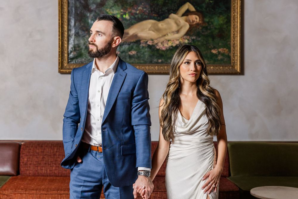 A couple in formal attire sits on a brown leather sofa in an elegant room with artwork on the walls.
