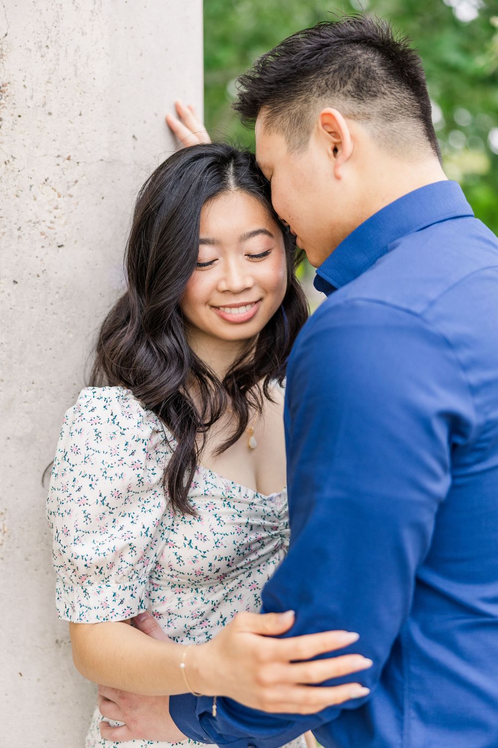 A couple in a blue suit and floral dress embrace lovingly against a light colored wall during an outdoor photo session.