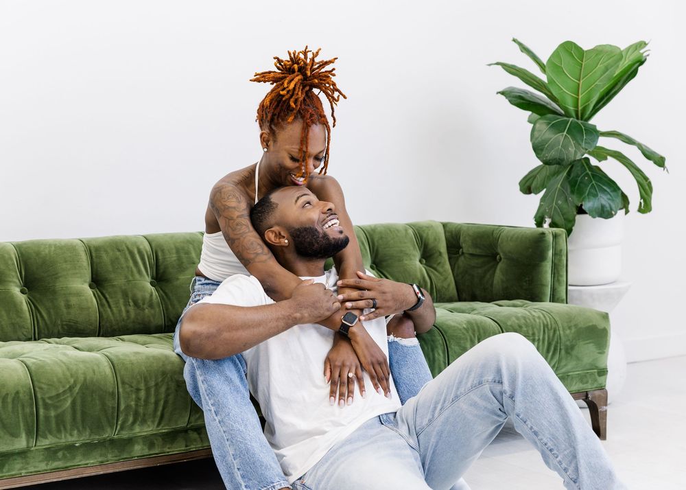 Two people share a playful moment while relaxing on a plush green velvet couch with a potted plant in the background.