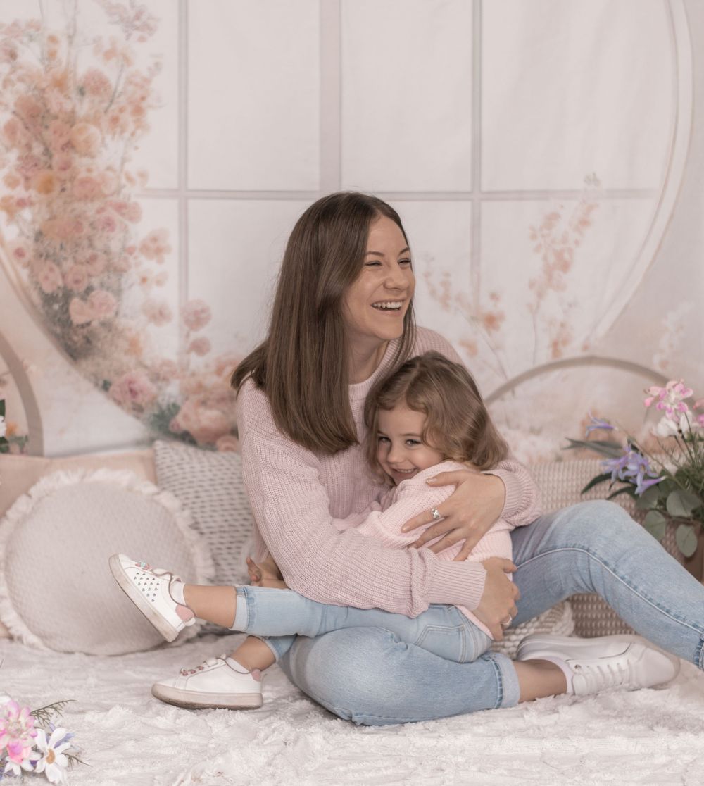 Mother and daughter lying on white bed with pink flowers and decorative accents in soft, cozy setting.