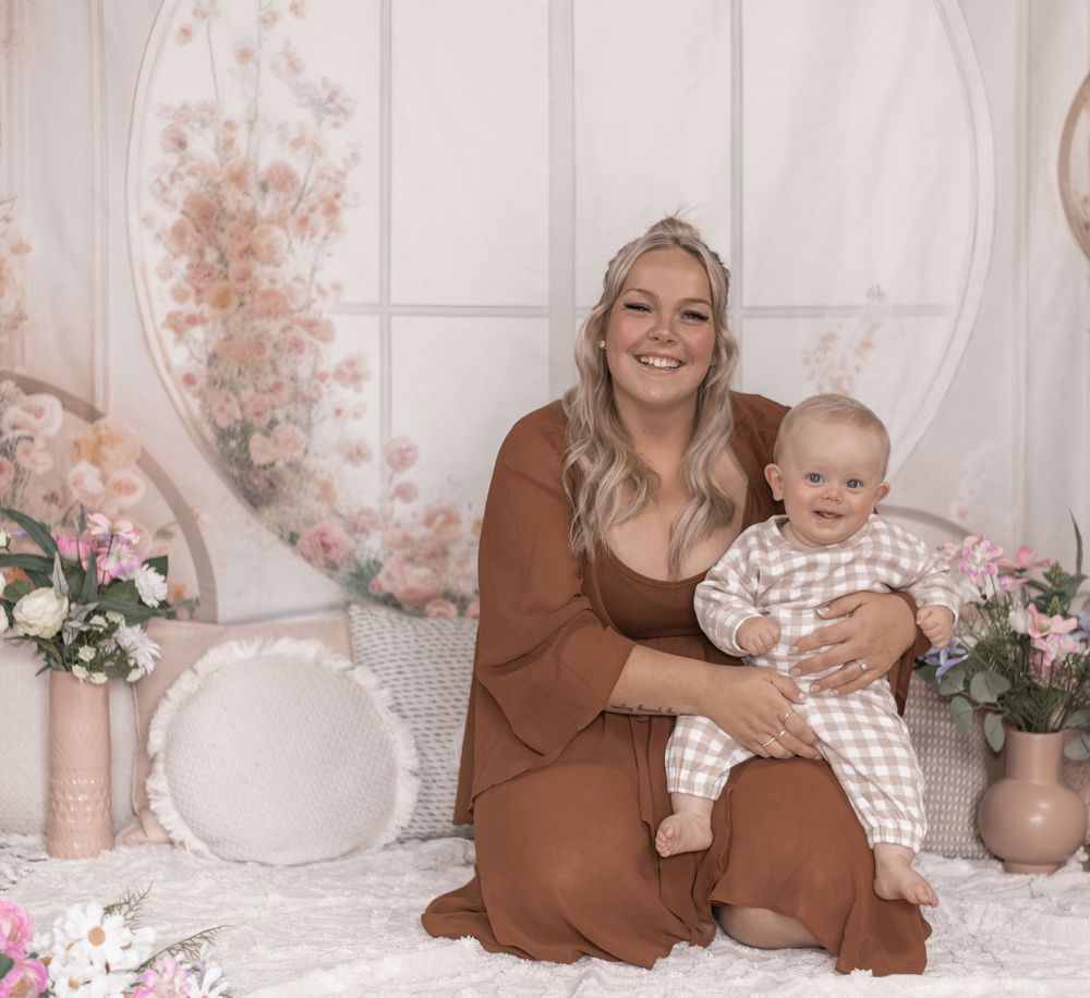 A parent and baby pose together on a decorative set with floral arrangements and soft pink accents.