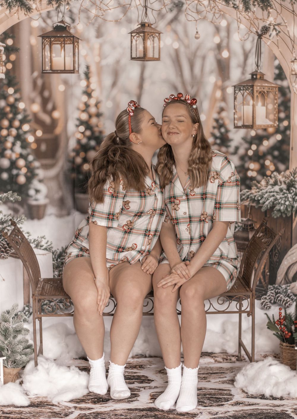 Two people in matching plaid outfits sit together on a bench surrounded by lanterns and holiday decor in a dreamy setting.